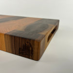 End grain cherry and walnut cutting board with a distinct striped pattern on a white background, corner angle with logo and handles visible
